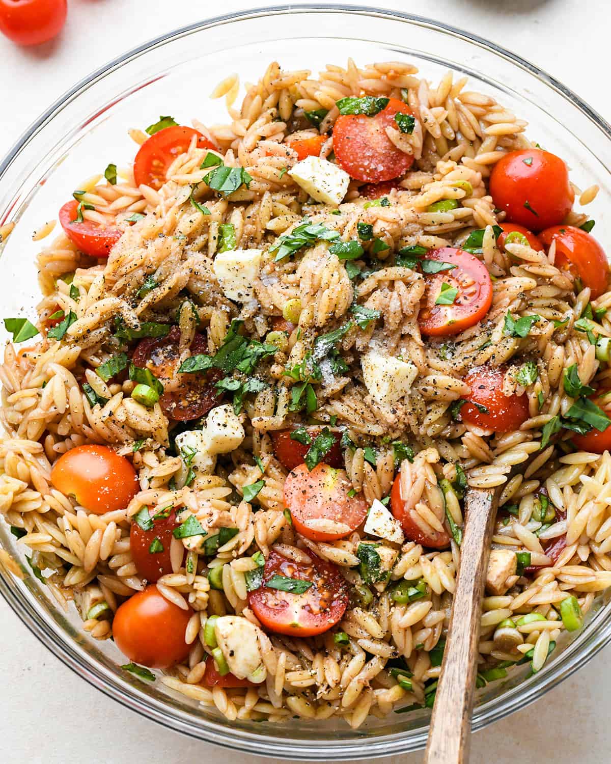 salt and pepper added to Caprese Orzo Salad in a bowl with a wooden stirring spoon