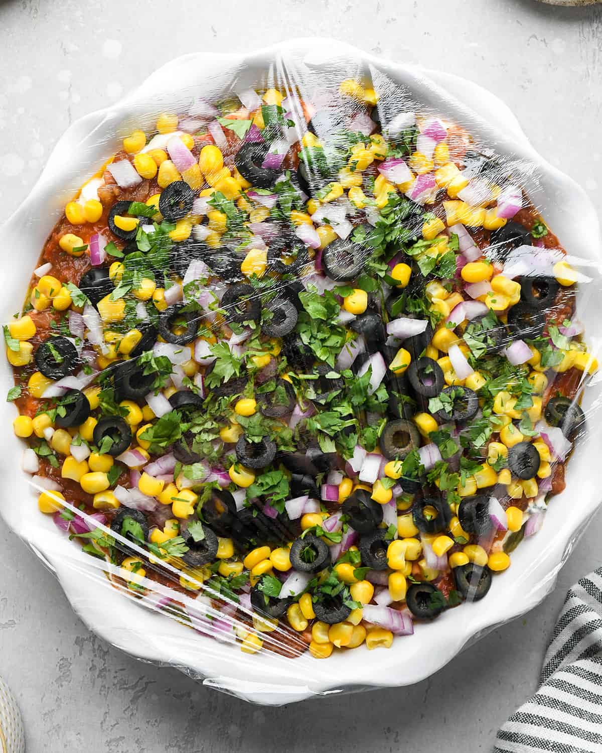 Mexican 7 Layer Dip in a pie dish covered in plastic wrap to chill