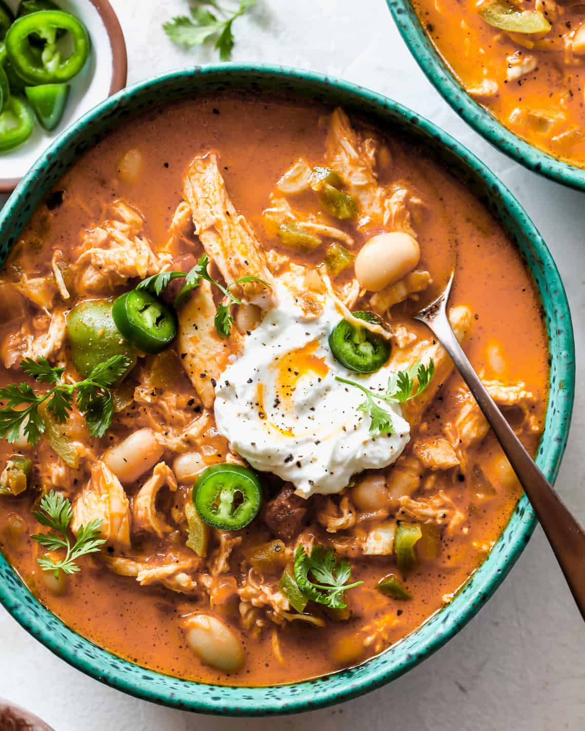 overhead view of a bowl of White Bean Chicken Chili garnished with sour cream, jalapeños, and cilantro with a spoon in it