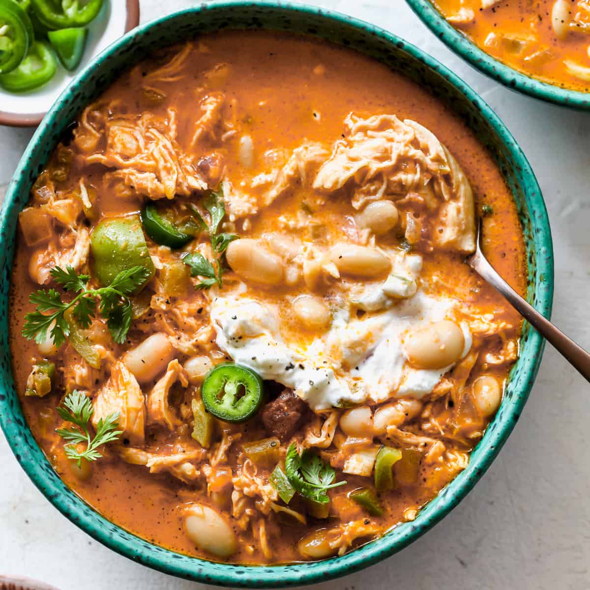 a bowl of White Bean Chicken Chili garnished with sour cream, jalapeños, and cilantro with a spoon in it